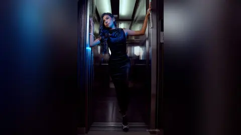 Elegant woman in a sleek black evening dress posing in a moody, dimly lit elevator.