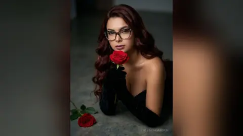 Elegant woman with glasses in a black dress holding a red rose in a moody, dark studio setting.