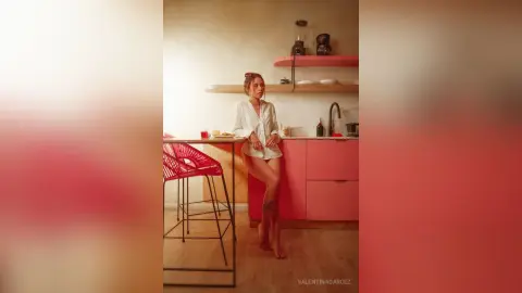 ValentinaGarcez stands in a kitchen wearing a white button-down shirt, showing her bare legs and leaning against a pink counter.