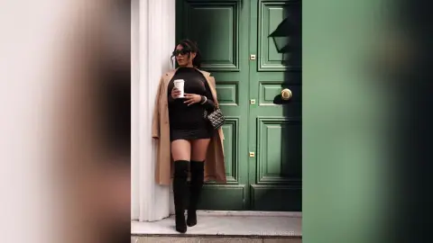 Stylish woman in a black mini dress and tall boots holding coffee by a green door in a chic urban setting.