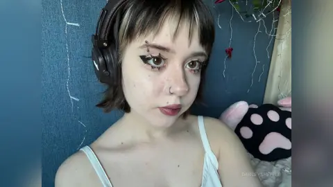 Young woman with dark bangs and star eyeliner wearing headphones in a cozy, moody bedroom setting.