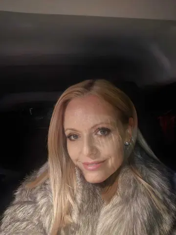 Smiling blonde woman in a cozy grey faux fur coat sitting in a car at night.