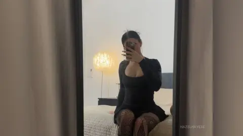 Young woman in a black long-sleeve mini dress taking a mirror selfie in a cozy, dimly lit bedroom.