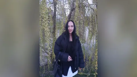 Young woman in a black puffer jacket standing in a misty willow forest with a serene mood.