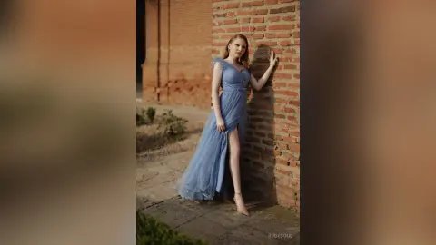 RubySoul poses against a brick wall wearing a long, light blue dress with a high slit showing her bare leg.
