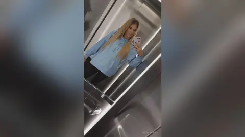 Blonde woman in a light blue hoodie taking a mirror selfie in a modern elevator.