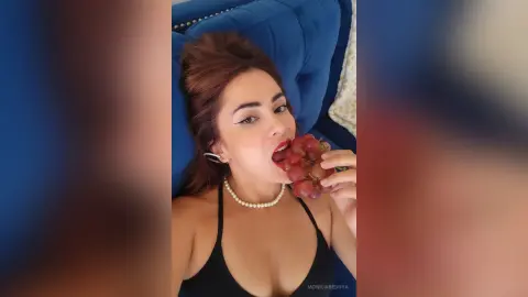 Beautiful woman in a black top and pearl necklace eating red grapes while relaxing on a blue velvet sofa.