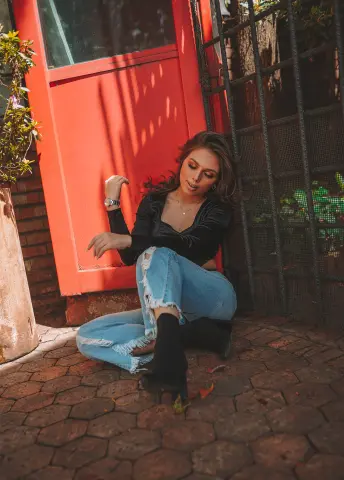 Stylish woman in black top and ripped jeans sitting on a brick patio by a red door in soft sunlight.