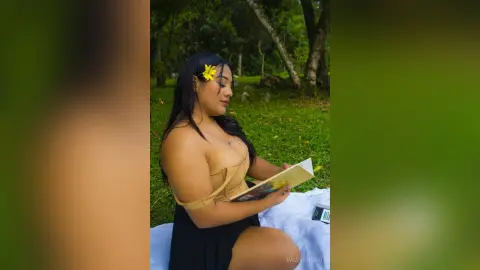 Young woman in a black dress reading a book on a white blanket in a lush green park setting.