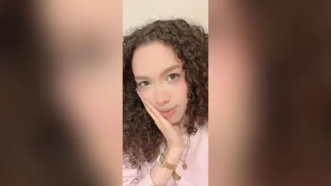 Young woman with curly brown hair wearing a pink top and gold watch in a soft, bright indoor setting.
