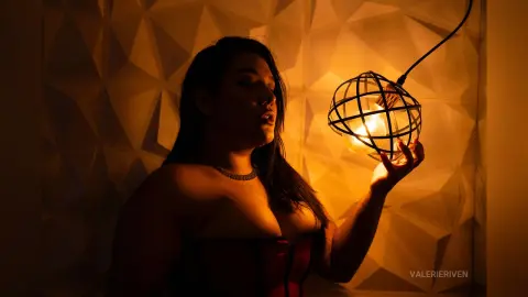 Moody silhouette of a woman in a red corset holding a glowing orb lamp against a geometric textured wall.
