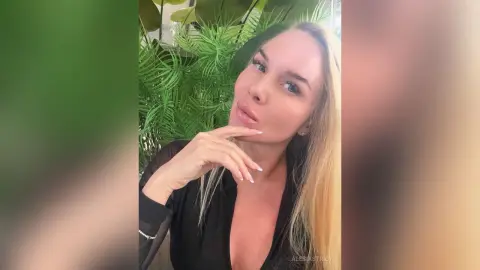 Blonde woman in a black top posing thoughtfully against a lush green plant background.