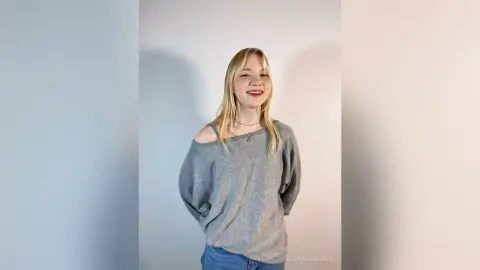 Smiling blonde woman in a grey off-the-shoulder sweater posing against a plain white background.