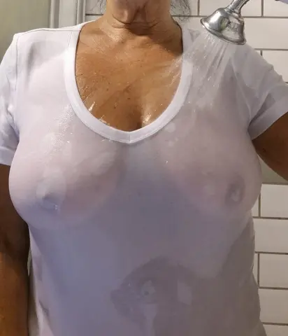 Woman in a wet white t-shirt standing under a shower head in a tiled bathroom.