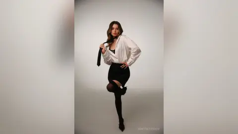 Stylish woman in a white button-down shirt and black skirt posing confidently against a minimalist studio background.