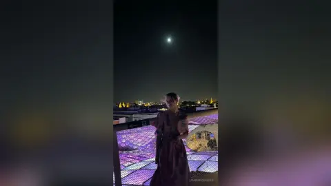LuanaRodgers stands on a rooftop at night under the moon, wearing a brown dress and looking toward the camera.