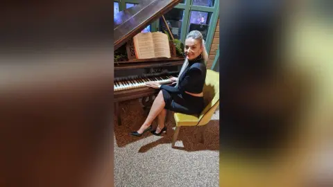 DelliaSmith sits on a yellow chair next to a dark wood piano, wearing a black long-sleeved dress and black high heels.
