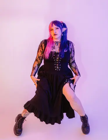 MagicalMae poses in a black lace dress and fishnet stockings against a pink background.