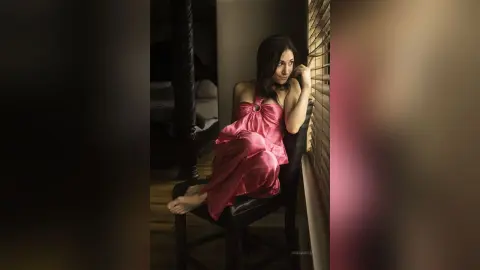 Melancholic woman in a pink satin dress sitting by a window in a dimly lit room.