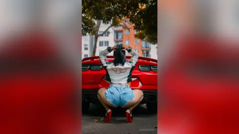 KimRoberth squats behind a red car, wearing light blue denim shorts and a white cropped jacket while holding her dark hair.