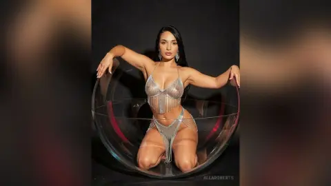 Glamorous woman in a sparkling silver sequin bikini sitting in a clear bubble chair against a black background.