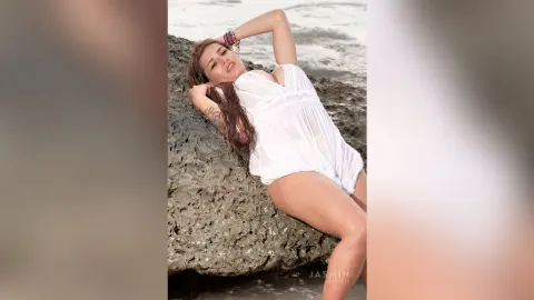 AndreaKalo poses against a large rock at the beach, wearing a short white lace-trimmed romper that shows off her legs and tanned skin.