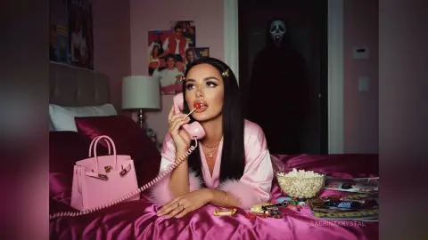 Woman in pink silk robe eating popcorn in a bedroom with Ghostface lurking in the background.