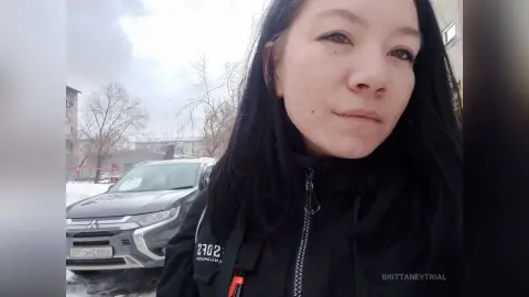 Young woman in a black winter jacket standing outdoors on a cloudy day near a parked silver SUV.
