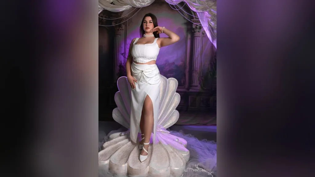 BettyBlossom — BettyBlossom poses in a white crop top and high-slit skirt while sitting on a large decorative seashell prop.