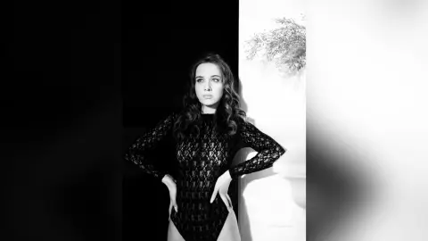 StacyHarland poses in a black lace long-sleeve bodysuit against a dark background.
