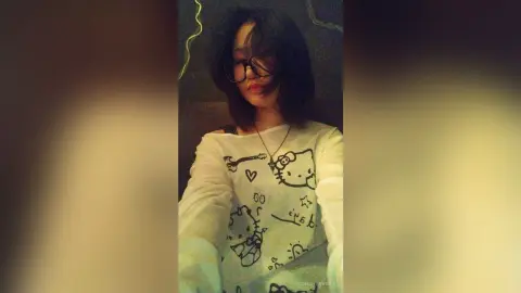TishaOyervides wears glasses and a white long-sleeve shirt with black Hello Kitty drawings, posing against a dark background.