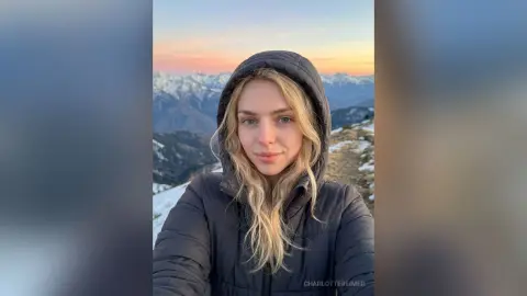 CharlotteReimer wearing a black puffer jacket and hood while posing outdoors against a snowy mountain sunset background.