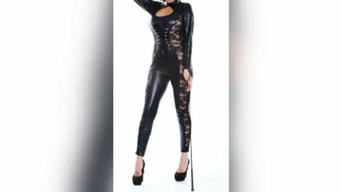 KateRichman stands in a black shiny latex bodysuit with lace cutouts, posing against a white background.