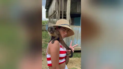 TefyRusso wears a red and white striped tank top and a straw hat while a small monkey sits on her shoulder in an outdoor setting.