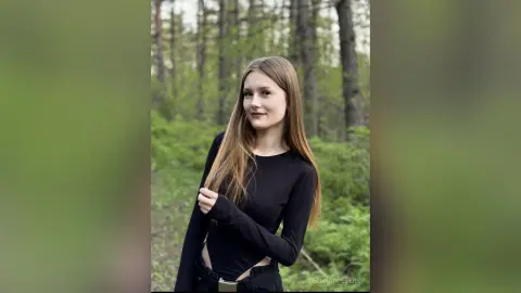 Young woman in a black long-sleeve bodysuit smiling in a lush green forest setting.