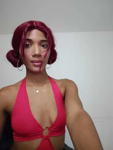 Young woman with red hair in a pink halter bikini top posing against a white wall.