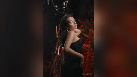 Elegant woman in a black strapless dress posing in a moody, dark floral studio setting.
