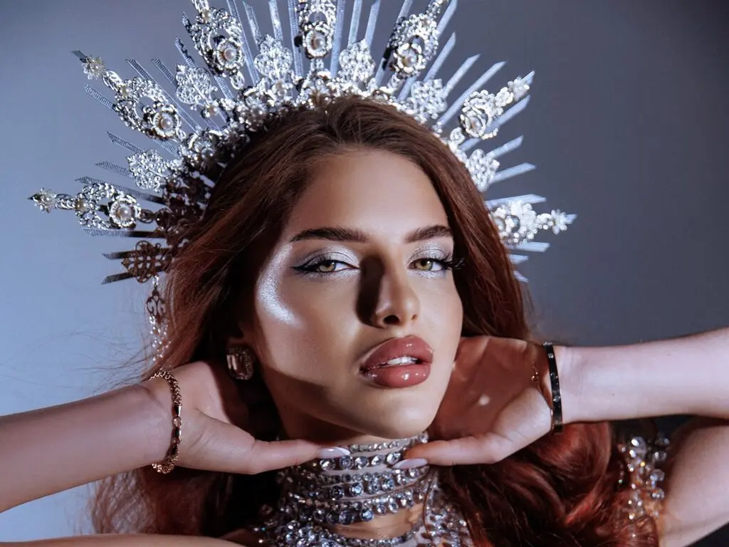 Glamorous woman in silver crown and sequined outfit posing with elegant makeup against a grey studio background.