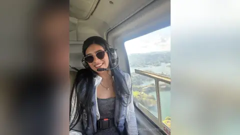 Smiling woman in sunglasses and headset enjoying a scenic helicopter flight over a coastal landscape.