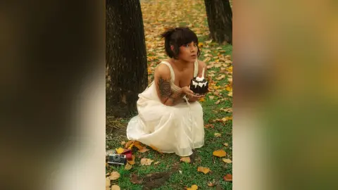 AlexiaVitali sits in a forest setting wearing a white dress and holding a small chocolate cake with a single candle.