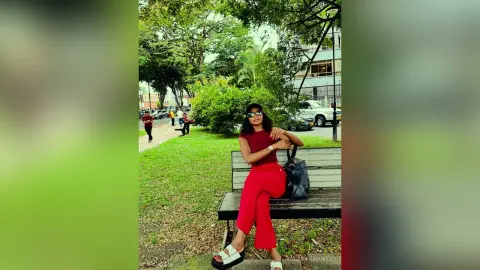 Young woman in a red top and trousers sitting on a park bench under lush green trees on a sunny day.