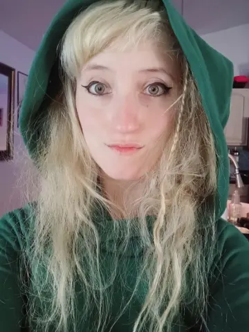 Blonde woman in a green hooded sweatshirt looking at the camera in a cozy indoor setting.