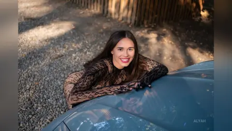 Smiling young woman in a black lace long-sleeve dress leaning on a car in a sunny outdoor setting.