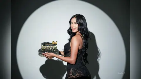 Aleja smiles while holding a small black cake topped with a gold crown, wearing a black lace dress against a white background.
