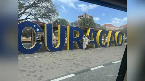 Tourist couple standing by the large blue and yellow Curacao sign on a sunny cobblestone street.