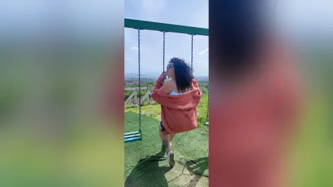 Curvy woman in a pink cardigan and denim shorts relaxing on a swing set against a scenic landscape view.