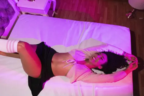 Smiling woman in a white crop top and black skirt relaxing on a bed under pink ambient lighting.