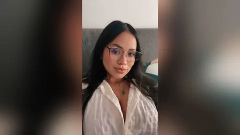 Young woman with long dark hair and glasses wearing a white linen shirt in a cozy indoor setting.