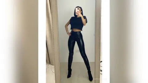 Stylish woman in a black crop top and tight leather pants taking a mirror selfie in a bright room.