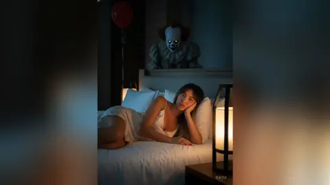 Katia lies on a bed in a white slip, looking sleepy in a dark room with a creepy clown watching her from the background.